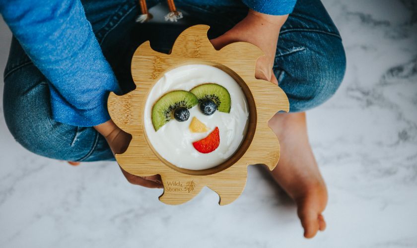 Child holding wooden weaning bowl filled with yogurt and fruit in the shape of a smiley face.